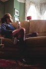 Comprimento total de homem pensativo hipster segurando telefone celular no sofá em casa — Fotografia de Stock