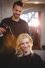 Портрет усміхненої жінки, яка сушить волосся феном у перукарні — стокове фото