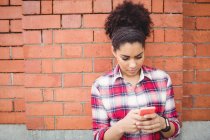 Junge Frau benutzt Handy, während sie sich an Ziegelmauer lehnt — Stockfoto