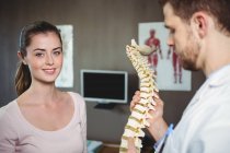 Fisioterapeuta sosteniendo modelo de columna vertebral mientras el paciente sonríe a la cámara en la clínica - foto de stock