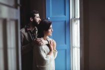 Чоловік обіймає жінку, дивлячись через вікно вдома — стокове фото
