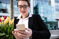Усміхнена молода бізнес-леді, використовуючи мобільний телефон проти офісної будівлі — стокове фото