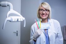 Retrato de dentista sorridente segurando três escovas de dentes na clínica odontológica — Fotografia de Stock