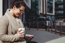 Улыбающаяся женщина с помощью цифрового планшета, держа одноразовую чашку на тротуаре — стоковое фото
