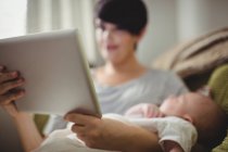Foyer sélectif de la mère en utilisant une tablette numérique pendant que le bébé dort dans son bras au salon — Photo de stock