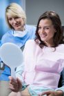 Улыбающаяся пациентка, смотрящая в зеркало с сидящей на кровати стоматологом — стоковое фото