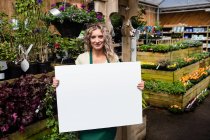 Retrato de uma florista feminina sorridente segurando um cartaz em branco no centro do jardim — Fotografia de Stock