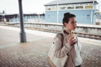 Молода жінка з напоєм дивиться на платформу залізничного вокзалу — стокове фото