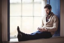 Mann benutzt Handy während er zu Hause auf Fensterbank sitzt — Stockfoto