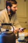 Bule e xícara em uma mesa em casa com o homem usando telefone no fundo — Fotografia de Stock