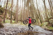 Side view of biker carrying bicycle in stream by trees at forest — Stock Photo