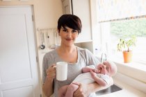 Ritratto di madre che tiene in braccio il suo bambino mentre prende una tazza di caffè in cucina — Foto stock