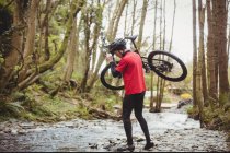 Mountain biker carrying bicycle while crossing stream in forest — Stock Photo