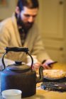 Teekanne und Tasse auf einem Tisch zu Hause mit Mann mit Telefon im Hintergrund — Stockfoto