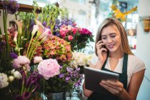 Floristería femenina usando tableta digital mientras habla por teléfono móvil en la tienda de flores - foto de stock