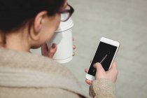Frau benutzt Handy während sie Einweg-Kaffeetasse in der Hand hält — Stockfoto