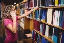 Frau telefoniert mit Handy, während sie Buch aus Bücherregal in Bibliothek entfernt — Stockfoto