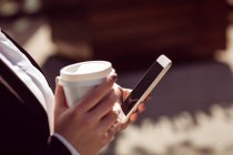 Imagem cortada de mulher segurando telefone celular e copo de café descartável — Fotografia de Stock