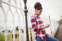 Seitenansicht einer jungen Frau, die ihr Handy benutzt, während sie sich an Geländer lehnt — Stockfoto