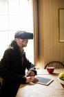 Sonriente hipster usando simulador de realidad virtual con tableta digital en la mesa en casa - foto de stock