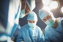 Cirujano mirando en cámara mientras sus colegas realizan la operación en la sala de operaciones - foto de stock