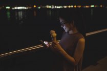 Young woman using mobile phone while having ice-cream at night — Stock Photo