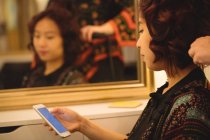 Mulher usando telefone celular ao obter cabelo endireitado no salão de cabeleireiro — Fotografia de Stock