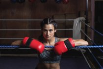 Tired female boxer in boxing gloves leaning on ropes of boxing ring at fitness studio — Stock Photo