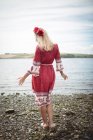 Вид сзади на беззаботную блондинку в красном платье и цветочную тиару, стоящую у реки — стоковое фото