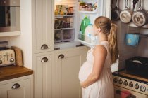 Беременная женщина ищет еду в холодильнике на кухне — стоковое фото