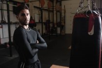 Retrato de boxeador de pie con los brazos cruzados al lado del saco de boxeo en gimnasio - foto de stock