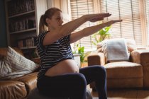 Mulher grávida realizando exercício de alongamento na bola de fitness na sala de estar em casa — Fotografia de Stock