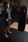 Retrato de boxeador de pé com as mãos nos quadris ao lado de saco de perfuração no estúdio de fitness — Fotografia de Stock