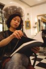 Mulher elegante ler uma revista enquanto sentado sob um secador de cabelo no salão de cabeleireiro — Fotografia de Stock