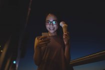 Junge Frau benutzt Handy, während sie nachts Eis isst — Stockfoto