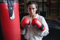 Portrait of woman in karate kimono and boxing gloves standing near punching bag in fitness studio — Stock Photo