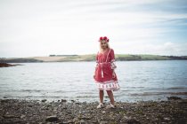 Atractiva mujer rubia en vestido rojo y tiara de flores de pie cerca del río y mirando a la cámara - foto de stock
