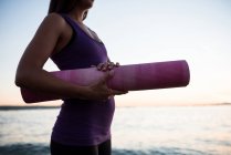 Средняя часть женщины, стоящей с ковриком для йоги на пляже в сумерках — стоковое фото