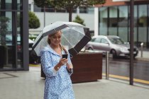 Mulher bonita segurando guarda-chuva ao usar o smartphone durante a estação chuvosa — Fotografia de Stock