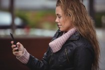 Vista lateral da mulher bonita usando jaqueta de couro e usando smartphone na rua — Fotografia de Stock