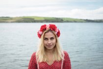 Carefree blonde woman in flower wreath looking at camera near river — Stock Photo