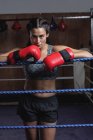 Boxeador cansado en guantes de boxeo apoyado en cuerdas de anillo de boxeo en el gimnasio y mirando a la cámara - foto de stock