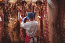 Carniceiro a colocar etiquetas nas carcaças de carne vermelha penduradas na arrecadação do talho — Fotografia de Stock