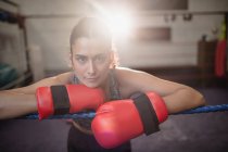 Retrato de boxeador feminino em luvas de boxe apoiado na corda do anel de boxe no estúdio de fitness, retroiluminado no fundo — Fotografia de Stock
