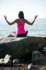 Visão traseira da mulher realizando ioga na rocha no dia ensolarado e mostrando gesto mudra — Fotografia de Stock