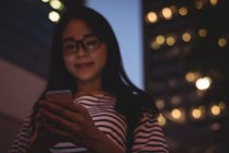 Mujer joven usando el teléfono móvil en la ciudad por la noche - foto de stock
