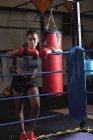 Портрет женщины-боксера в боксёрских перчатках, опирающейся на канат для бокса в фитнес-студии — стоковое фото