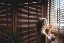 Femme réfléchie regardant par la fenêtre dans le salon à la maison — Photo de stock