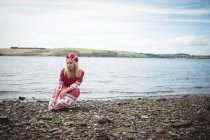 Donna bionda spensierata in abito rosso e fiore tiara seduta vicino al fiume — Foto stock