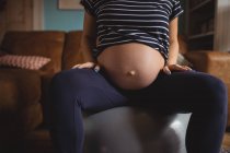 Мидсекция беременной женщины, сидящей на мяче в гостиной дома — стоковое фото